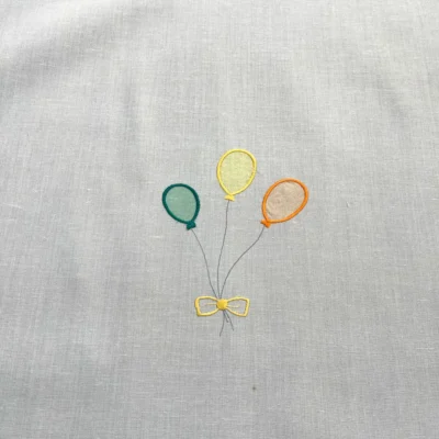 Primo piano di una tendina a vetro bianca con ricamo a forma di tre palloncini colorati (verde, giallo, arancione) e fiocco.