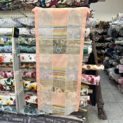 Tendina a vetro con motivo geometrico rosa pesca e grigio esposta in negozio su rotoli di tessuto